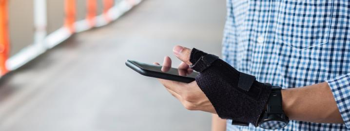 Een man met een blauwgeruit hemd houdt met een hand in een brace - Un homme vêtu d'une chemise à carreaux bleus tient un smartphone d'une main dans une attelle et parcourt l'écran